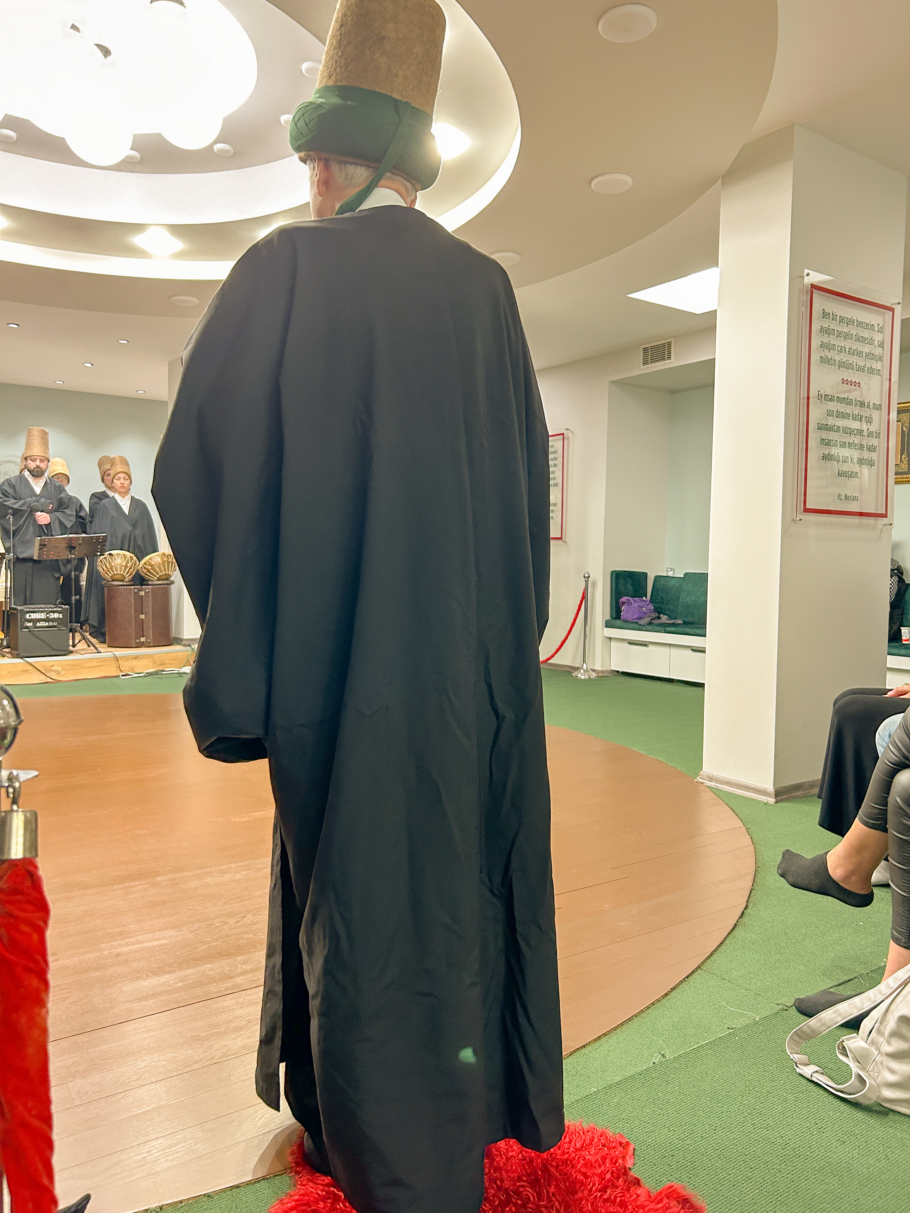 Sheikh standing on top of a red sheepskin rug. The sheikh is dressed in a black robe with a brown and green hat.