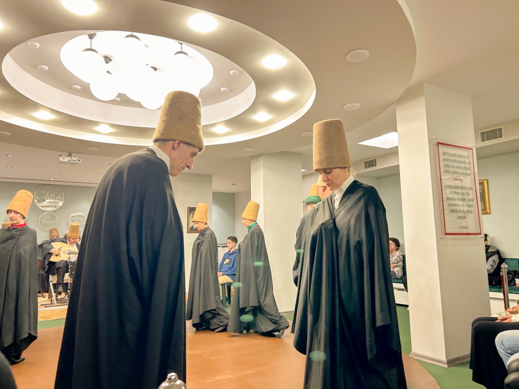 Two participants facing each other and bowing. Both are wearing black robes with tall, brown, cylindrical hats. 