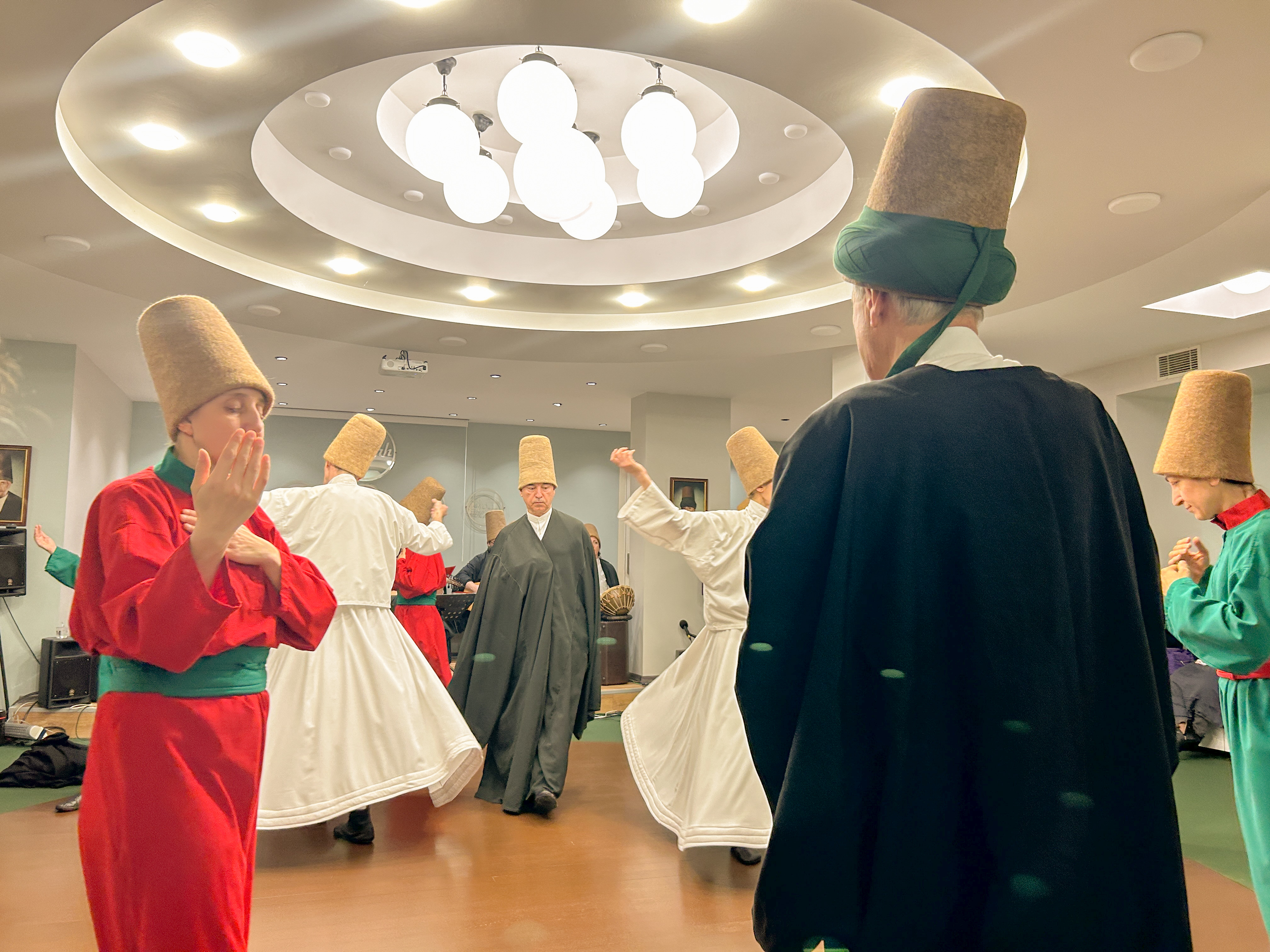 Room with two participants, whirling dervishes, wearing all white and spinning. Other participants wearing red and green move in a circle around them. Sheikh is standing and watching.