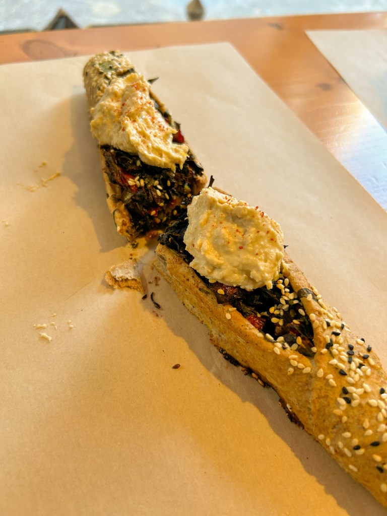 Baguette shaped bread with green vegetable filling and dollops of hummus on top. Sesame seeds are sprinkled on top.