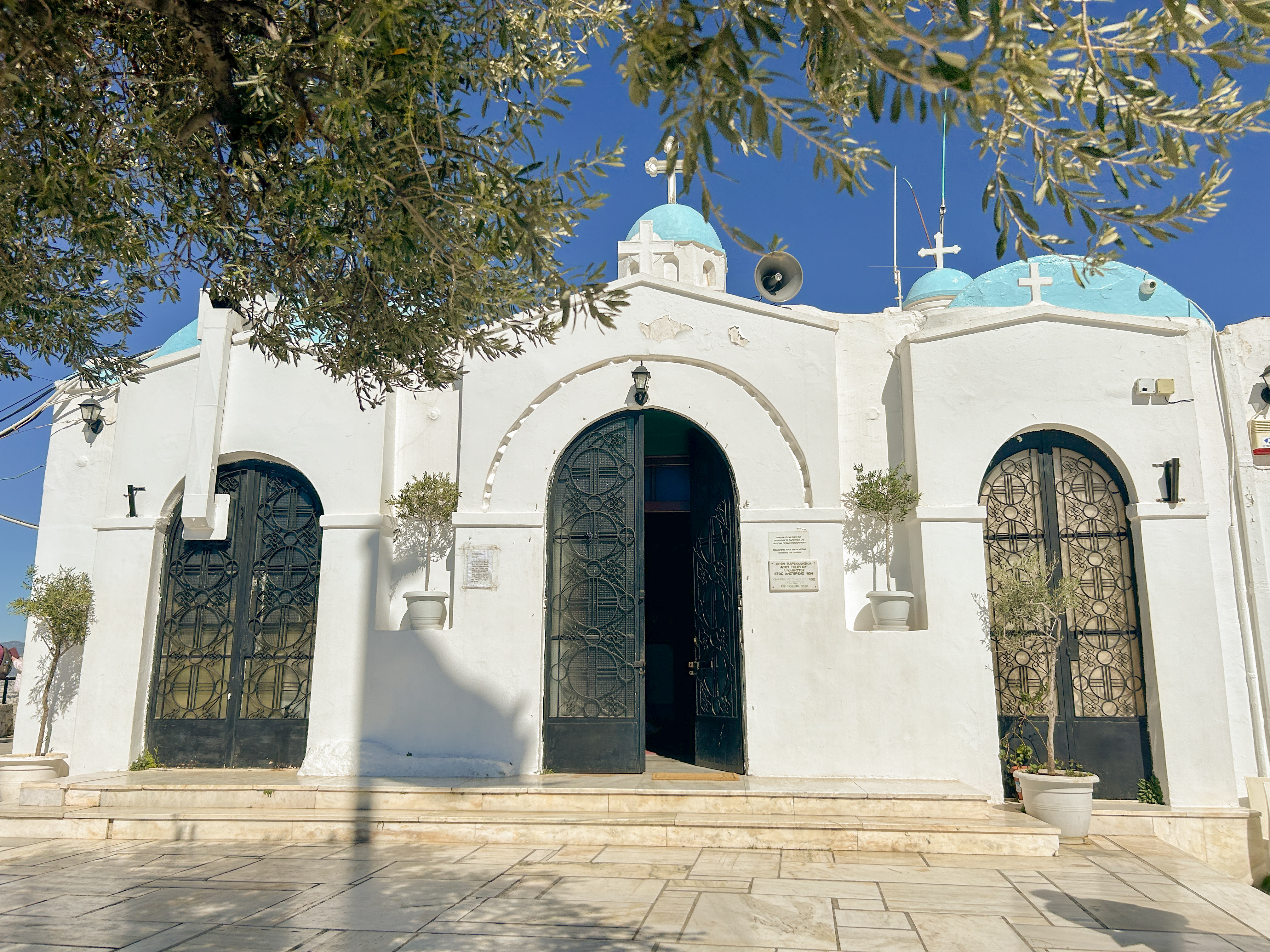 White and turquoise blue church at the top of the mountain. One iron gate is open. Several white crosses are atop the church.