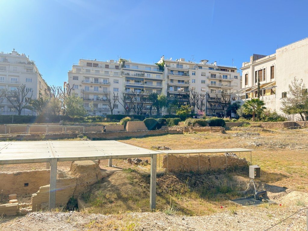 Flat park surrounded by apartment buildings. Inside the park are small ruins in the ground covered by awnings. 