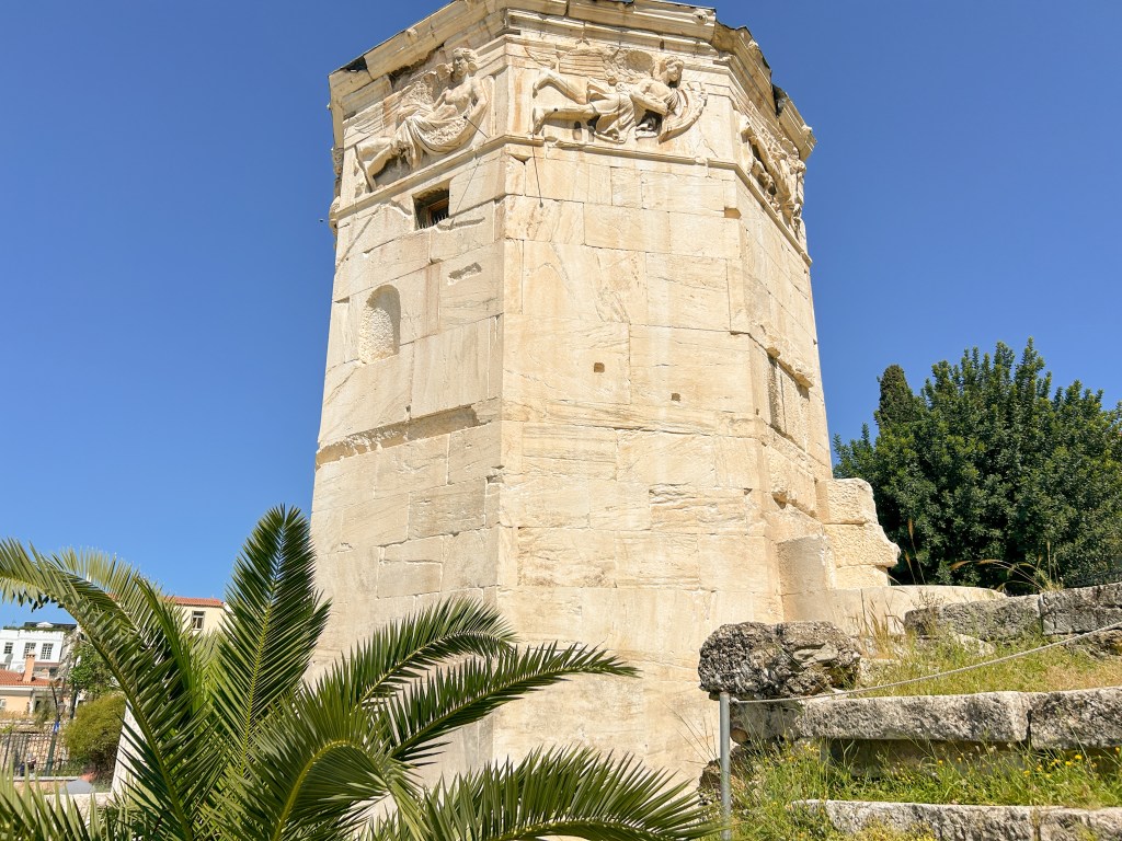 Tower of Winds made of marble stands tall with carvings on the top. A palm tree is partially in the foreground.