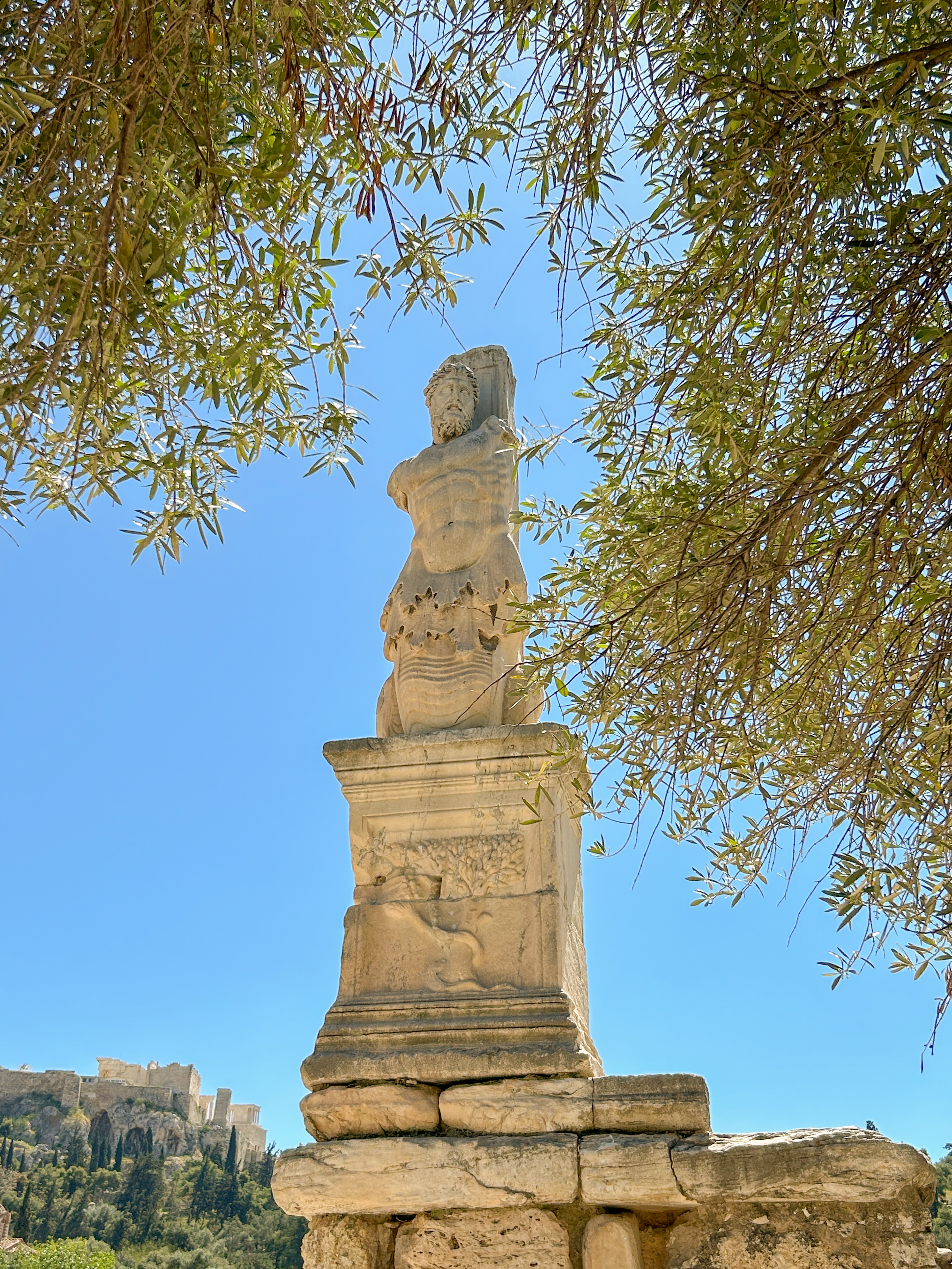 Marble statue of a man sits atop a gate. The acropolis is visible in the background.