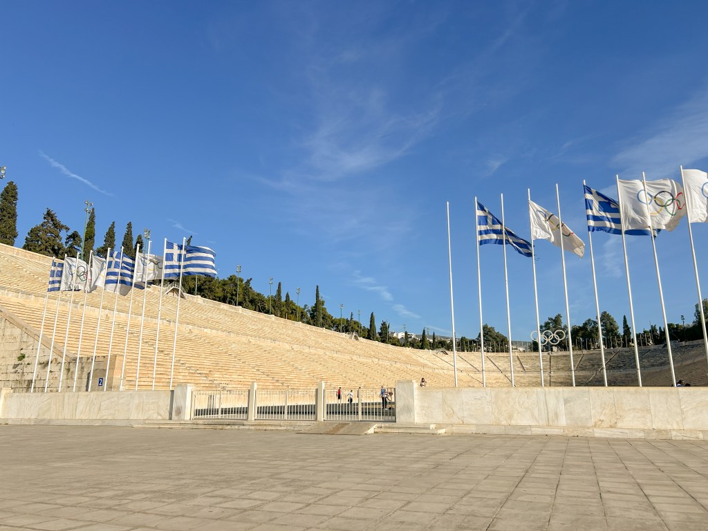Glistening marble stadium in a horse shoe shape. Many Greek and olympic flags stand in a row and wave in the wind.