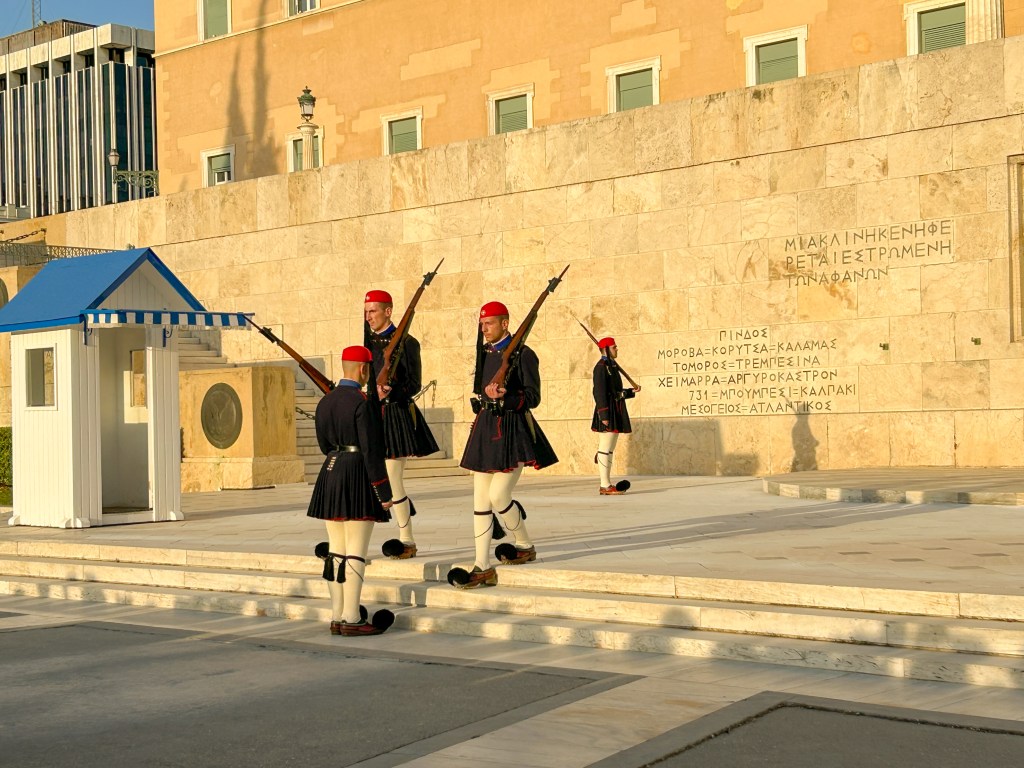 Four guards in navy blue uniforms with white leggings and curvy shoes with pom poms. Soldier are carrying guns and changing the guard.