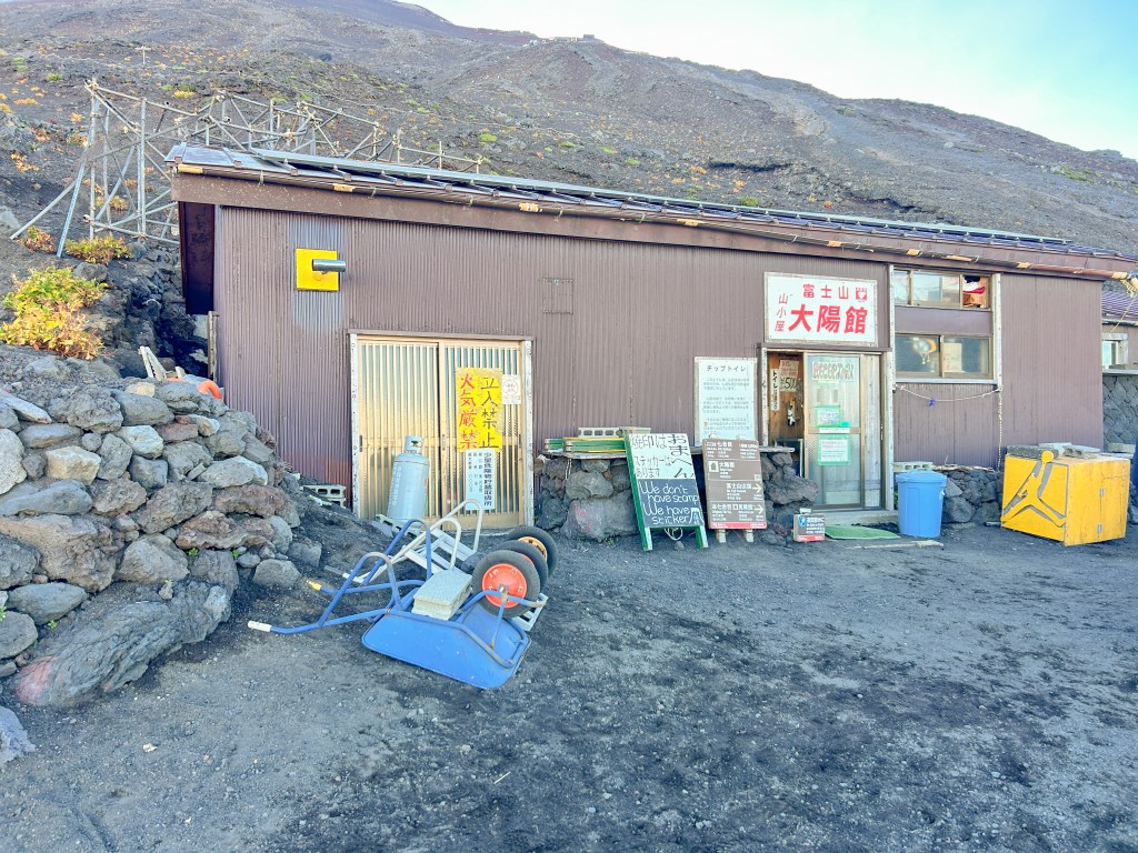 Brown rectangular shed with sliding doors and signs in Japanese language outside. The environment is rocky and sparse. 